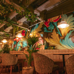 A restaurant interior featuring bold, colourful botanical wall art, hanging plants, cushioned seating, and warm pendant lighting.