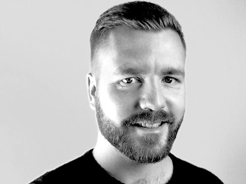 A black-and-white portrait of a person wearing a dark crew-neck shirt. The image is cropped to show the head and upper shoulders, with a plain light background. The person has short, neatly styled hair.