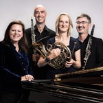 Kathryn Stott and members of Ensemble 360 (who are holding their musical instruments) stand behind a grand piano, smiling at the camera.