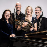 Kathryn Stott and members of Ensemble 360 (who are holding their musical instruments) stand behind a grand piano, smiling at the camera.
