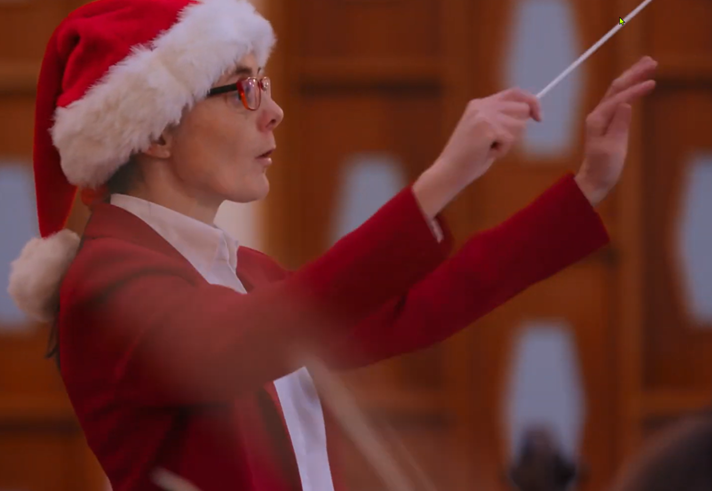 A woman in a red outfit and a Santa hat conducts an orchestra.