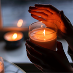 A close-up of a pair of hands holding a candle in a glass pot.