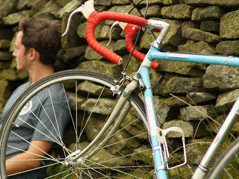 A cyclist sat leaning against a wall having a rest, out in the countryside, with his bike propped up next to him.