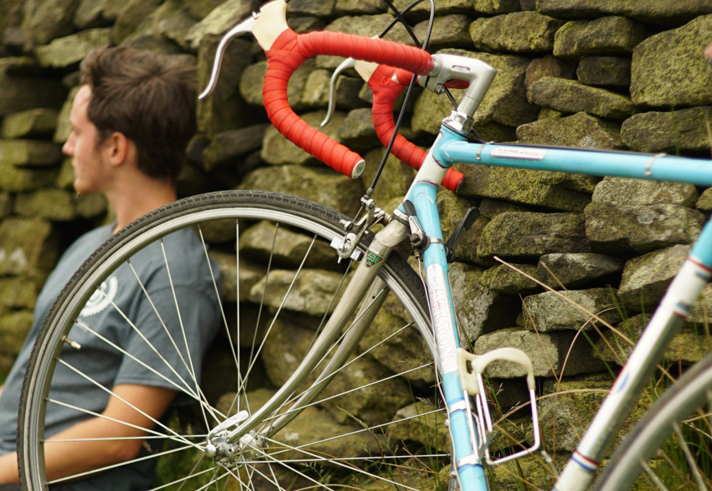 A cyclist sat leaning against a wall having a rest, out in the countryside, with his bike propped up next to him.