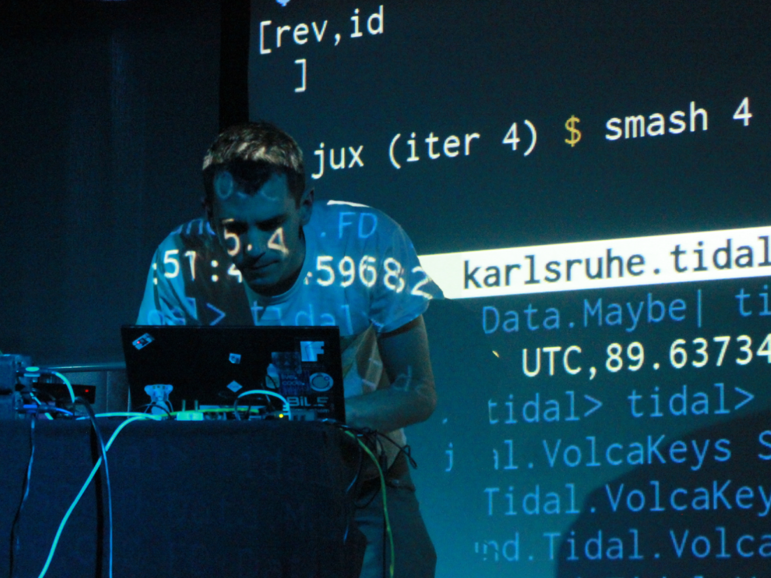 A person stands at a laptop during a live coding performance, illuminated by blue light as lines of programming code are projected across the screen and their body. Cables and audio equipment sit on the table while the projected text creates a dynamic, tech-focused atmosphere.