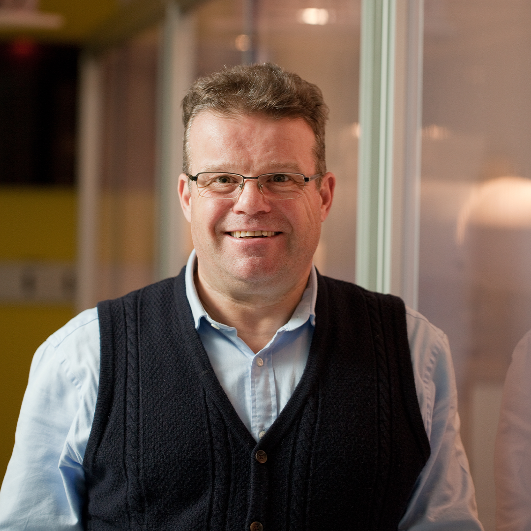 A person wearing a light blue button-up shirt layered with a dark textured vest, standing indoors near a glass partition. The background includes softly lit panels and a warm-toned interior, suggesting an office or professional setting.