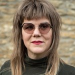 Person with long, layered, light brown hair featuring darker roots and lighter tips, wearing a dark turtleneck top, standing in front of a textured stone wall background.