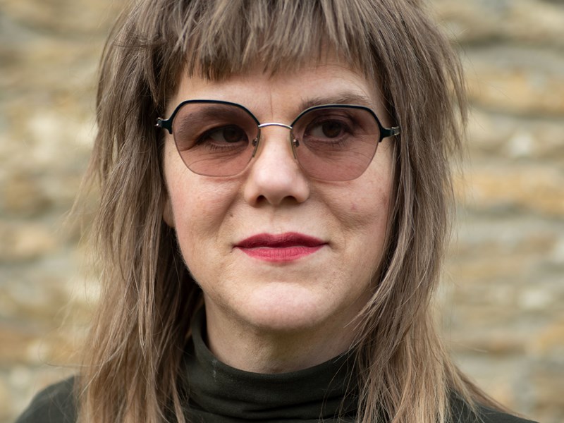 Person with long, layered, light brown hair featuring darker roots and lighter tips, wearing a dark turtleneck top, standing in front of a textured stone wall background.