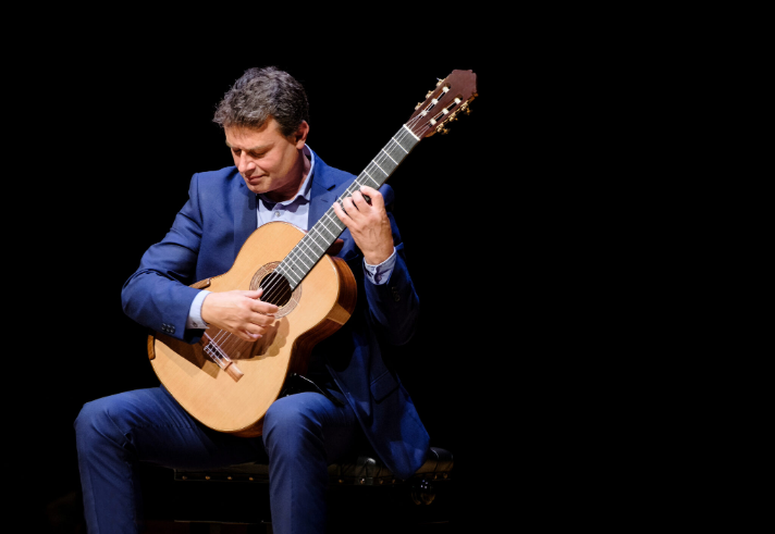 A person wearing a dark blue suit is seated on a chair, playing a classical guitar. The guitar has a light wooden body with nylon strings, and the musician’s hands are positioned on the fretboard and soundhole, suggesting a performance. The background is completely black, drawing focus to the player and the instrument.