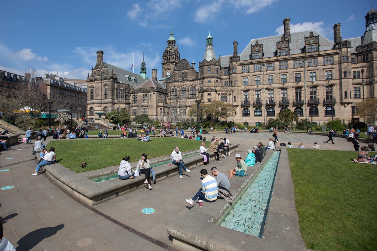 People are sitting and relaxing in an open public square with green lawns and paved walkways. The area features a long, narrow water feature with turquoise tiles, and groups of individuals are scattered across benches and grass. In the background stands a large historic building with ornate architecture, multiple towers, and green copper roofs under a bright blue sky with scattered clouds.