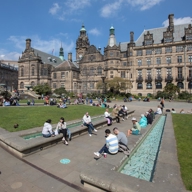 People are sitting and relaxing in an open public square with green lawns and paved walkways. The area features a long, narrow water feature with turquoise tiles, and groups of individuals are scattered across benches and grass. In the background stands a large historic building with ornate architecture, multiple towers, and green copper roofs under a bright blue sky with scattered clouds.