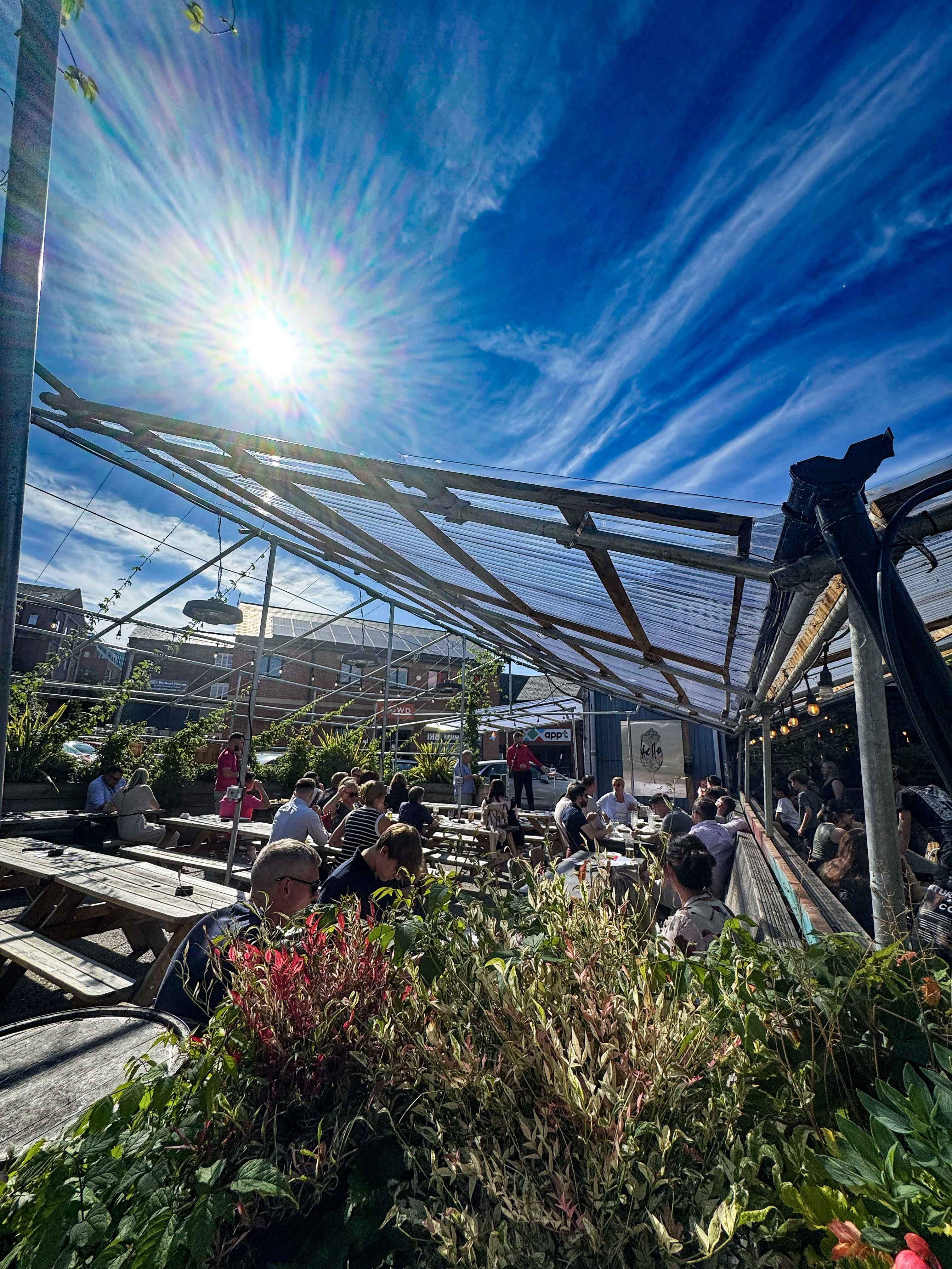 A vibrant outdoor beer garden packed with people at wooden benches beneath a blue-tinted canopy. Tall city buildings rise in the background, and plants and greenery decorate the space.