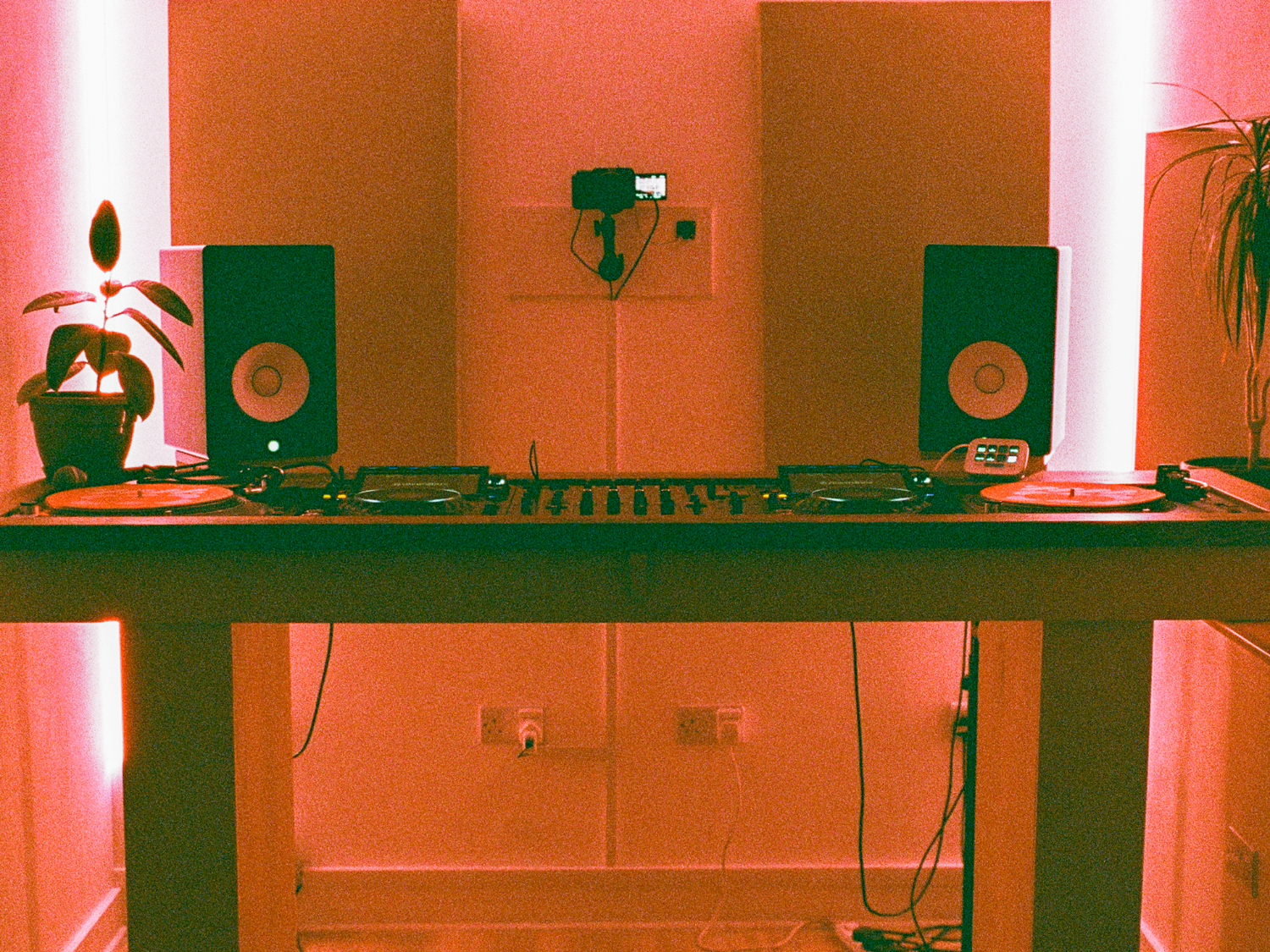 DJ setup in a studio with warm pink lighting, featuring a wooden table holding two turntables, a central mixer, and two black speakers with white cones. A small digital display sits on the right turntable. A potted plant is placed on the left side of the table, and another plant is visible on the right edge. Acoustic panels are mounted on the wall behind, with a small camera fixed above the table. Cables run beneath the table to wall sockets.