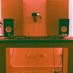 DJ setup in a studio with warm pink lighting, featuring a wooden table holding two turntables, a central mixer, and two black speakers with white cones. A small digital display sits on the right turntable. A potted plant is placed on the left side of the table, and another plant is visible on the right edge. Acoustic panels are mounted on the wall behind, with a small camera fixed above the table. Cables run beneath the table to wall sockets.
