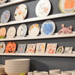 Hand-painted ceramic plates, tiles, mugs, and bowls displayed on wall-mounted shelves inside a pottery painting studio.