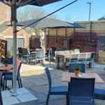 An outdoor pub terrace with wicker chairs, wooden tables and large parasols, surrounded by brick walls and planters on a sunny day.