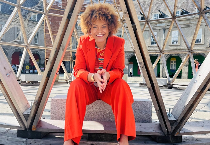 A woman in an orange suit sits, smiling, on a sculpture in the sunshine.
