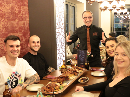 A group of people seated around a wooden dining table in a restaurant, with plates, drinks, and a large platter of assorted grilled meats and sides in the center. The table is set with menus, cutlery, and condiments. A person standing at the head of the table is wearing a black shirt with a brown tie and gesturing with both hands. The background includes decorative wall panels, a window, and a chandelier providing warm lighting.