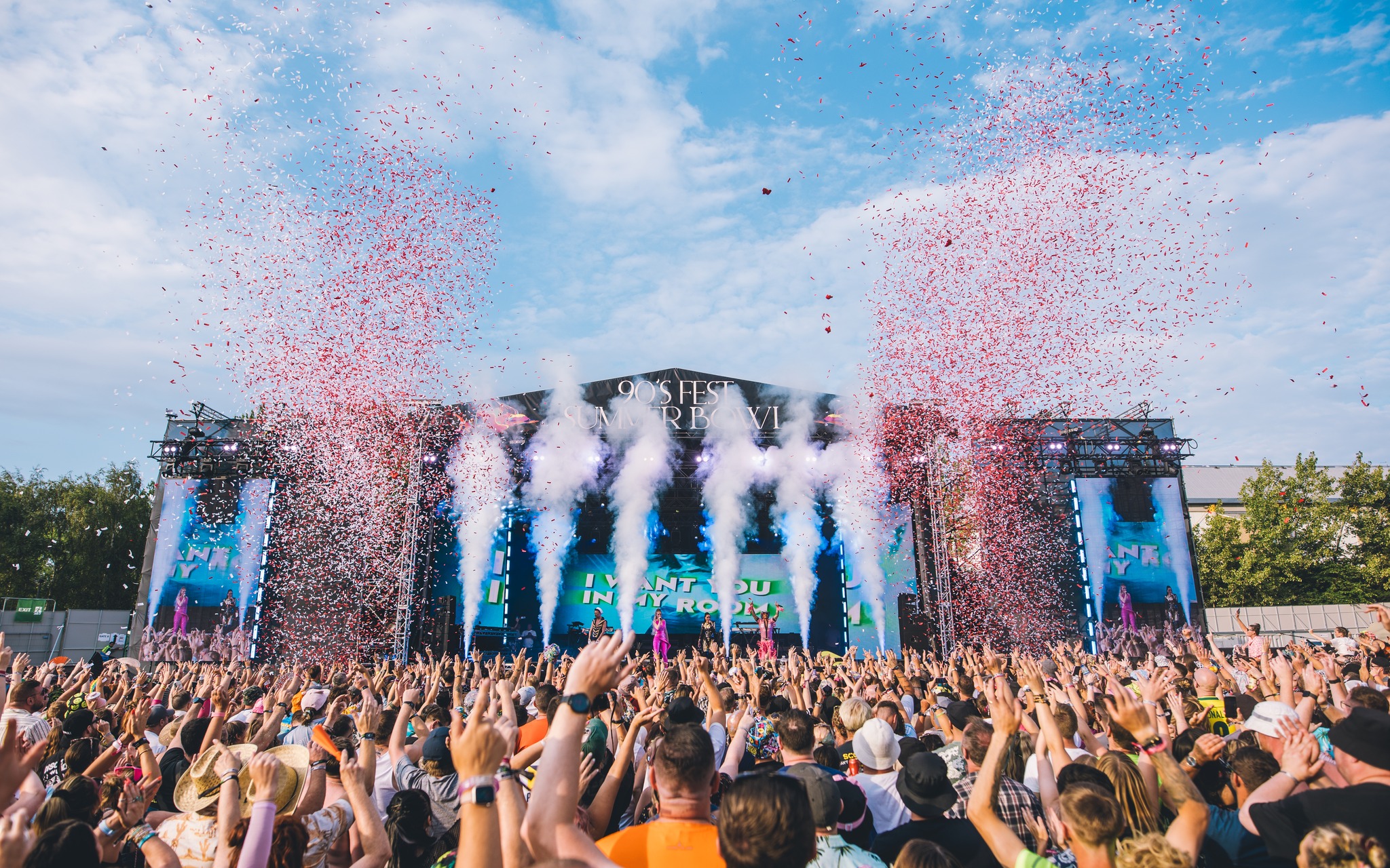 A packed audience at an outdoor music festival cheers in front of the “90’s Fest Summer Bowl” stage as large bursts of white smoke and red confetti erupt into the sky. The stage displays vibrant visuals, and the atmosphere is festive and colorful.
