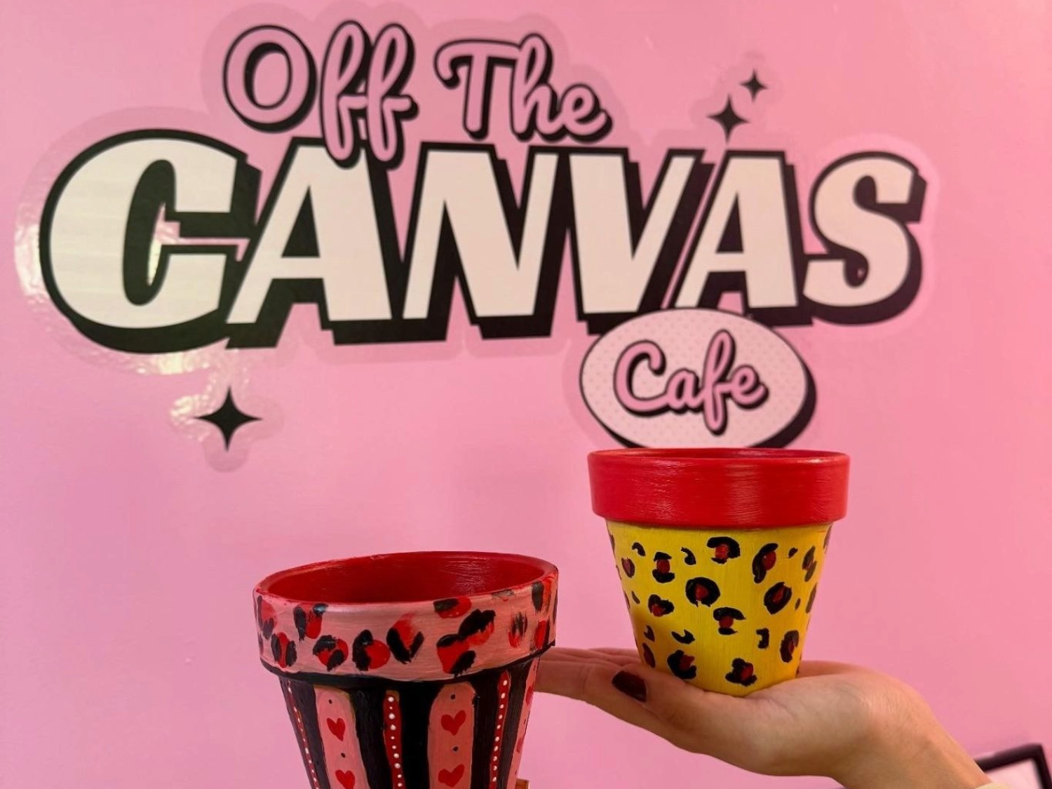 A hand is holding up a decorated pot in front of a  pink wall that has logo on it that reads 'OFF THE CANVAS'.