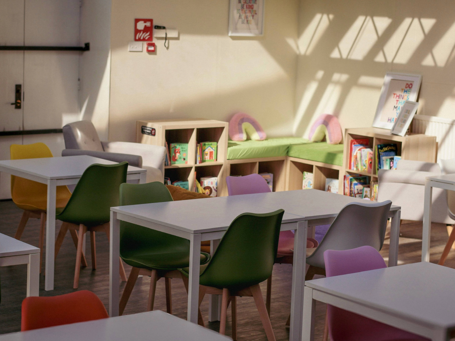 Bright, modern classroom or reading area with white tables and colourful chairs in shades of green, yellow, pink, orange, and grey. At the back, wooden shelves hold children’s books, and a green cushioned bench with pink pillows sits in the corner. Sunlight streams through large windows, casting shadows on the cream walls. A framed poster and a fire alarm are mounted on the wall above the shelves.