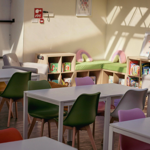 Bright, modern classroom or reading area with white tables and colourful chairs in shades of green, yellow, pink, orange, and grey. At the back, wooden shelves hold children’s books, and a green cushioned bench with pink pillows sits in the corner. Sunlight streams through large windows, casting shadows on the cream walls. A framed poster and a fire alarm are mounted on the wall above the shelves.
