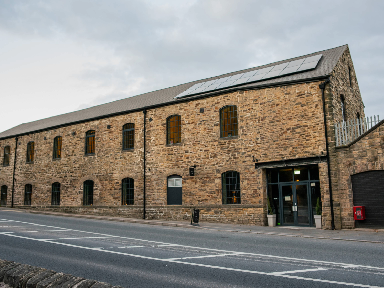 Exterior view of a large stone building with tall arched windows and a pitched roof fitted with solar panels. The structure has an industrial yet modern look, with a glass entrance door flanked by potted plants. It sits alongside a paved road with white markings, and the sky above is overcast. The building appears to be a converted mill or warehouse with contemporary features.