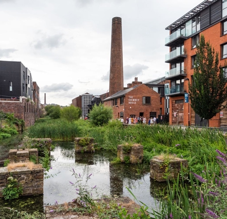 A scenic view of a canal surrounded by greenery and historic architecture. The water reflects the sky and nearby plants, with remnants of old stone structures partially submerged. On the right, modern red-brick and glass buildings line the canal, while a tall industrial chimney rises prominently in the background. A brick building labelled “The Chimney House” is visible near the centre, and purple wildflowers grow along the water’s edge.