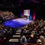 A packed theatre with tiered seating surrounding a central stage where two people are seated for a live talk under colourful lighting.