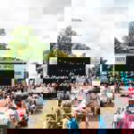 Outdoor event with a large stage featuring a choir performance, rows of deck chairs filled with spectators, and colourful bunting overhead. Inflatable structures and trees surround the area under a partly cloudy sky.