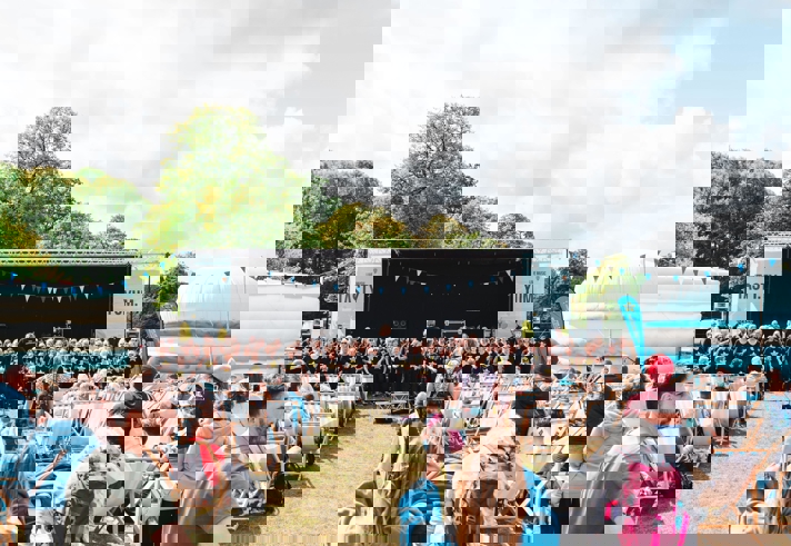 Outdoor event with a large stage featuring a choir performance, rows of deck chairs filled with spectators, and colourful bunting overhead. Inflatable structures and trees surround the area under a partly cloudy sky.