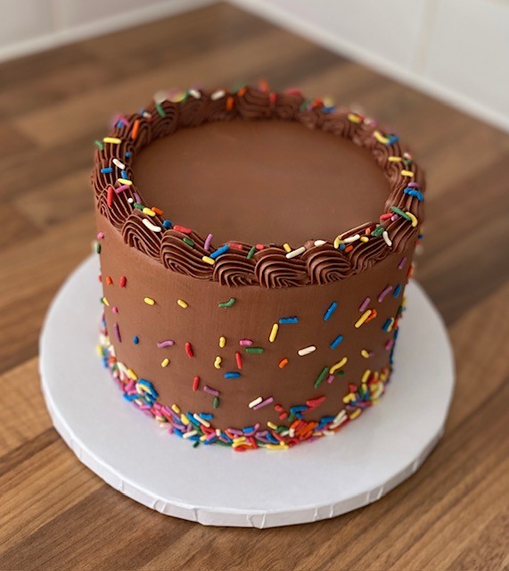 A round vegan chocolate cake covered in smooth chocolate frosting, decorated with colourful rainbow sprinkles around the sides and top edge. The cake sits on a white cake board on a wooden surface.