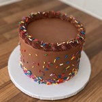A round vegan chocolate cake covered in smooth chocolate frosting, decorated with colourful rainbow sprinkles around the sides and top edge. The cake sits on a white cake board on a wooden surface.