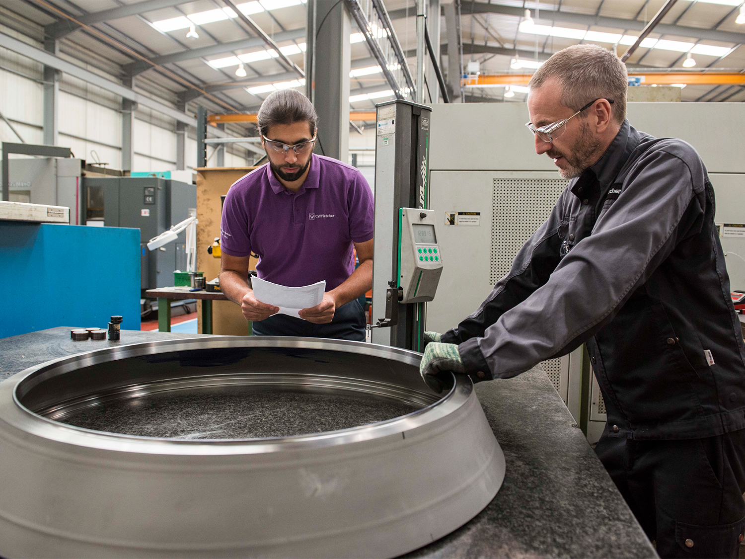 Two men working in an industrial setting. Both are wearing safety glasses and one of them is reading a sheet of paper.