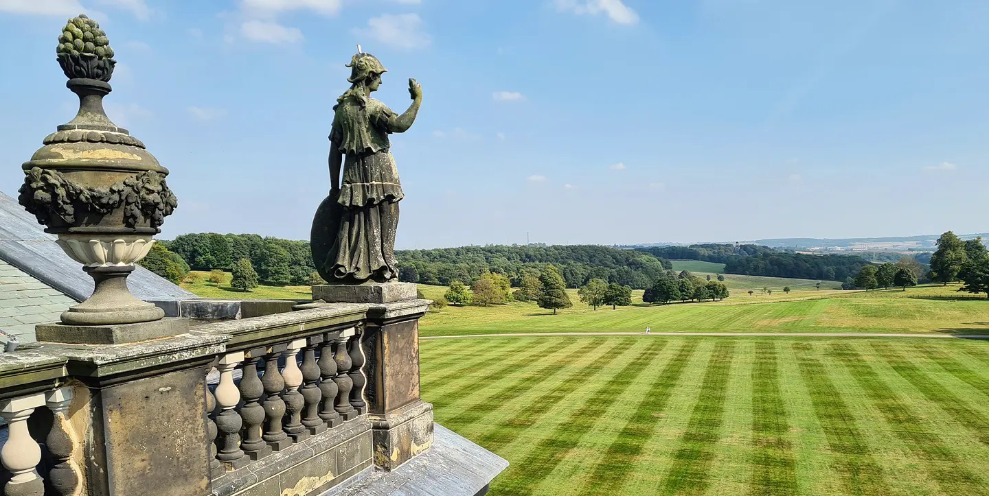 A stone terrace with a classical statue and ornate urn overlooks a vast, neatly striped lawn. The landscape stretches into the distance with clusters of trees, rolling hills and a pale blue sky with scattered clouds. Provide your feedback on BizChat
