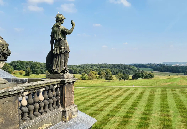 A stone terrace with a classical statue and ornate urn overlooks a vast, neatly striped lawn. The landscape stretches into the distance with clusters of trees, rolling hills and a pale blue sky with scattered clouds. Provide your feedback on BizChat