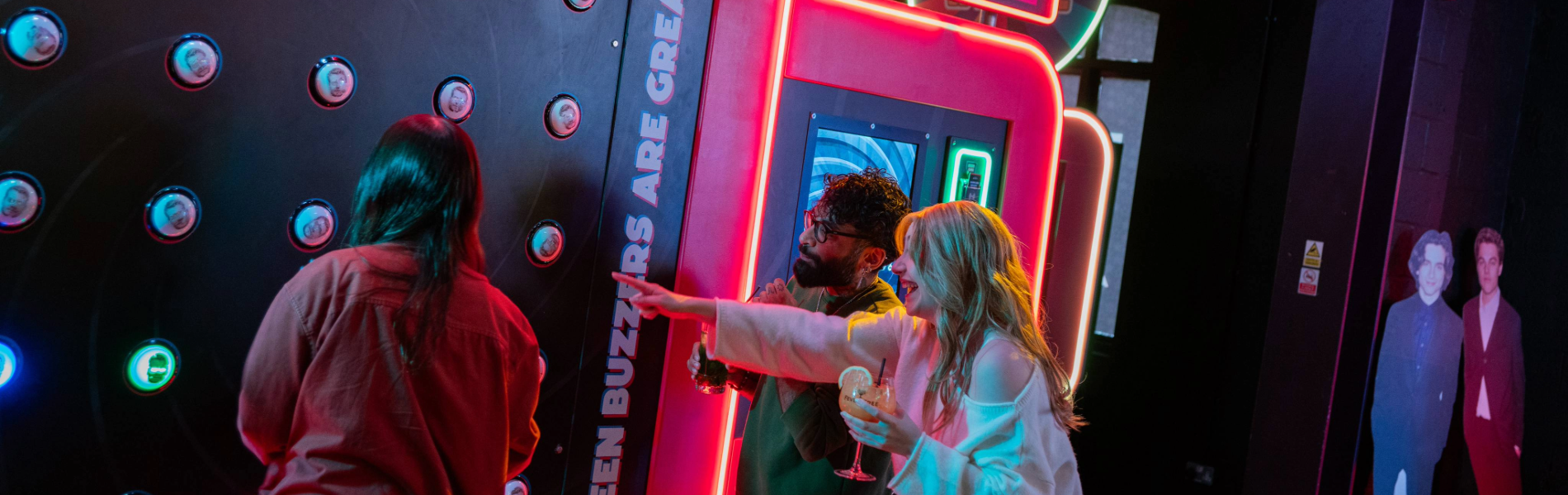 Three people playing an interactive arcade game called “Buzzer Beater” in a neon-lit venue. The game features a large vertical panel with multiple round buzzers and a digital score display reading “0550.” Bright signage on the machine includes phrases like “Green buzzers are great.” One person is actively playing, while another points at the panel, holding a drink. The setting is vibrant with colourful lights and a lively atmosphere.
