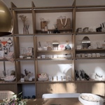 A pottery studio interior with wooden shelving filled with handmade ceramics, small homeware items, mirrors, and dried flowers, with a large table in the foreground set with ceramic plates.