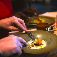 Close‑up of diners enjoying a plated fine‑dining dish at a restaurant table, with food being eaten using cutlery.