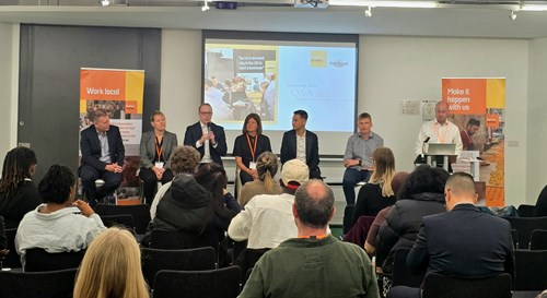 A panel of six speakers seated at the front of a conference room during a Q&A session at a Business Sheffield event. One speaker holds a microphone while others listen. The presentation screen behind them displays “Q&A Session,” with two orange Business Sheffield banners on either side reading “Work local” and “Make it happen with us.” Audience members are seated and engaged in the discussion.