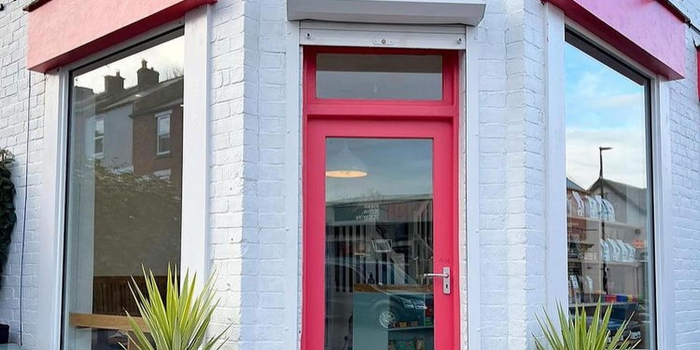 A brick building that has been painted white, with red window frames and door. There is a hand painted sign, in red, above the door that reads 'Corner Store'. The letter 'o' in the word Store is a smiley face.