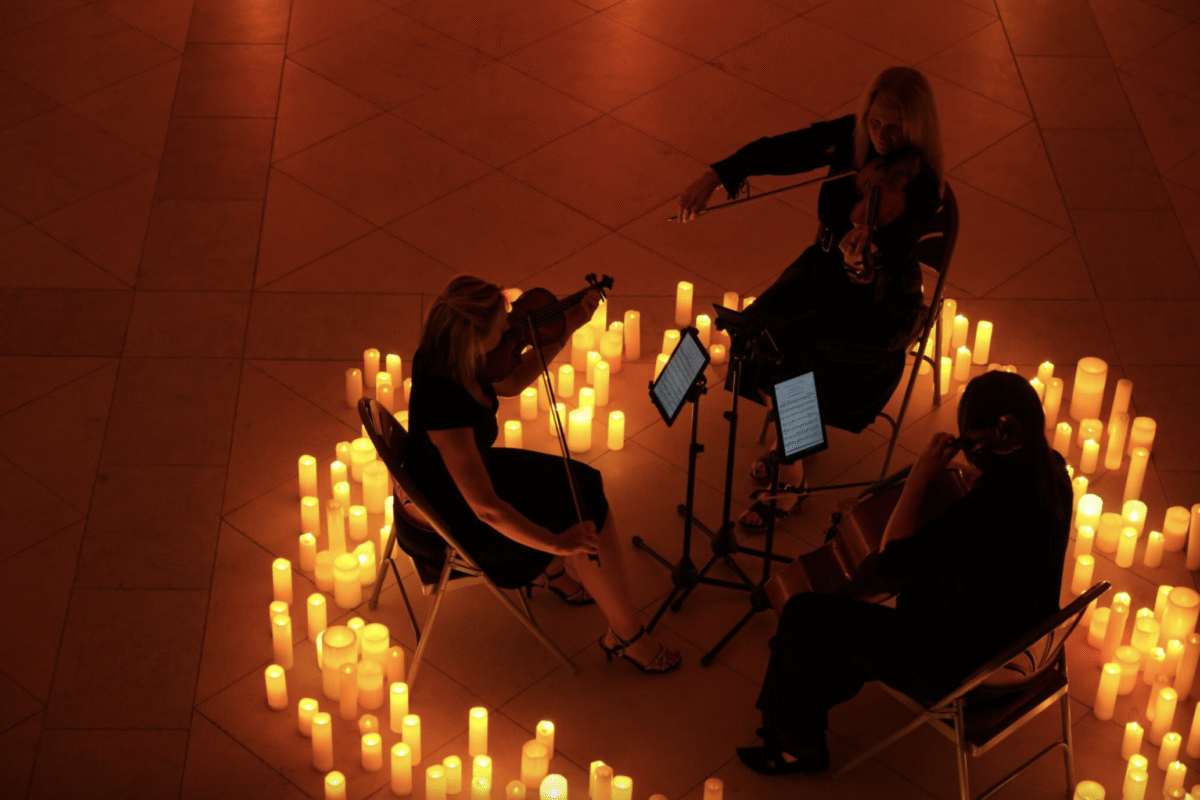 Three musicians seated in a circle perform with string instruments, surrounded by numerous glowing candles arranged on the floor. Music stands with sheet music are positioned in front of each player. The setting is dimly lit, creating a warm, intimate atmosphere with soft orange light reflecting off the tiled floor.