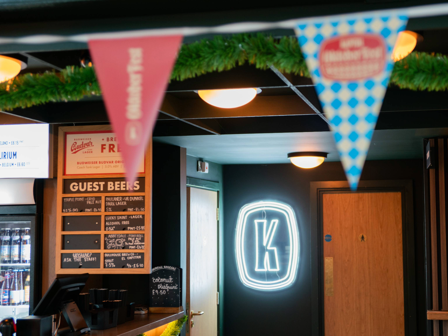 Interior of a beer hall featuring a bar counter with a chalkboard-style menu listing guest beers and prices. A neon sign with the letter “K” is illuminated on a dark wall near a wooden door. Colourful triangular bunting hangs across the ceiling, and green garland decorates the space. The setting has warm lighting and a modern, welcoming atmosphere.