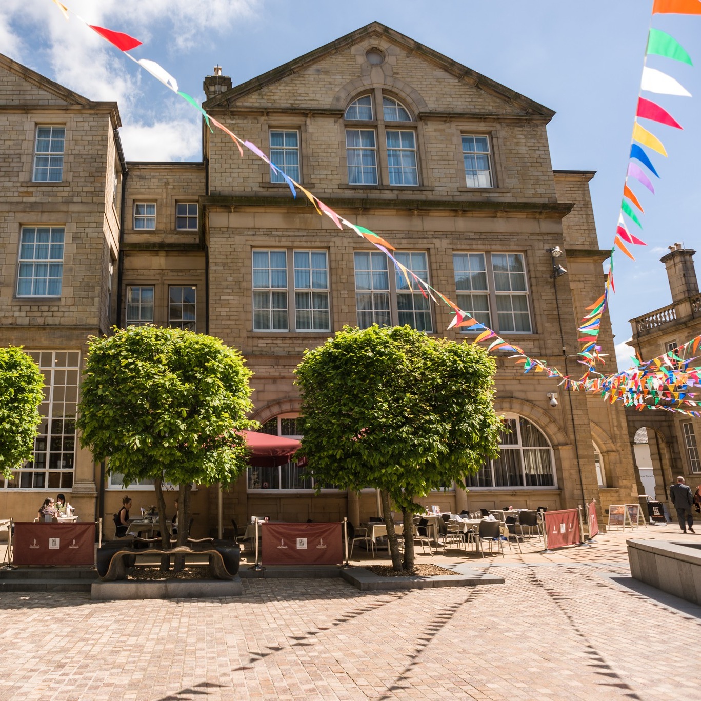 Courtyard with cobblestone paving in front of a historic stone building featuring large windows and a triangular roofline. Two neatly trimmed trees stand in the center, with outdoor seating and red barriers beneath them. Colorful triangular bunting is strung across the space under a bright blue sky with scattered clouds.