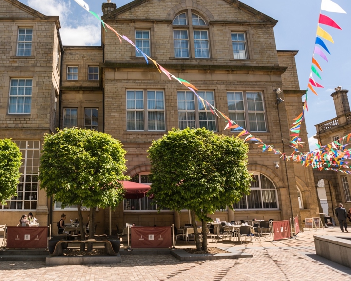 Courtyard with cobblestone paving in front of a historic stone building featuring large windows and a triangular roofline. Two neatly trimmed trees stand in the center, with outdoor seating and red barriers beneath them. Colorful triangular bunting is strung across the space under a bright blue sky with scattered clouds.