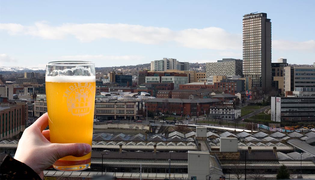 A hand holding a pint of beer up to the skyline of Sheffield from behind the train station