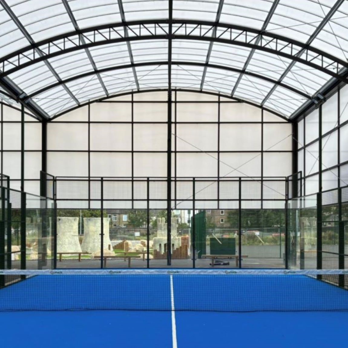 View of a covered padel court with a bright blue playing surface and a black net in the center. The structure has a high, arched roof made of translucent panels supported by metal beams, allowing natural light inside. Glass walls surround the court, and outdoor scenery is visible through them.