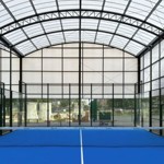 View of a covered padel court with a bright blue playing surface and a black net in the center. The structure has a high, arched roof made of translucent panels supported by metal beams, allowing natural light inside. Glass walls surround the court, and outdoor scenery is visible through them.