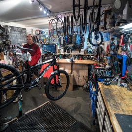 A photograph of the interior of The Bike Garage with the walls covered in tools and spare parts. There are two men working on bikes.