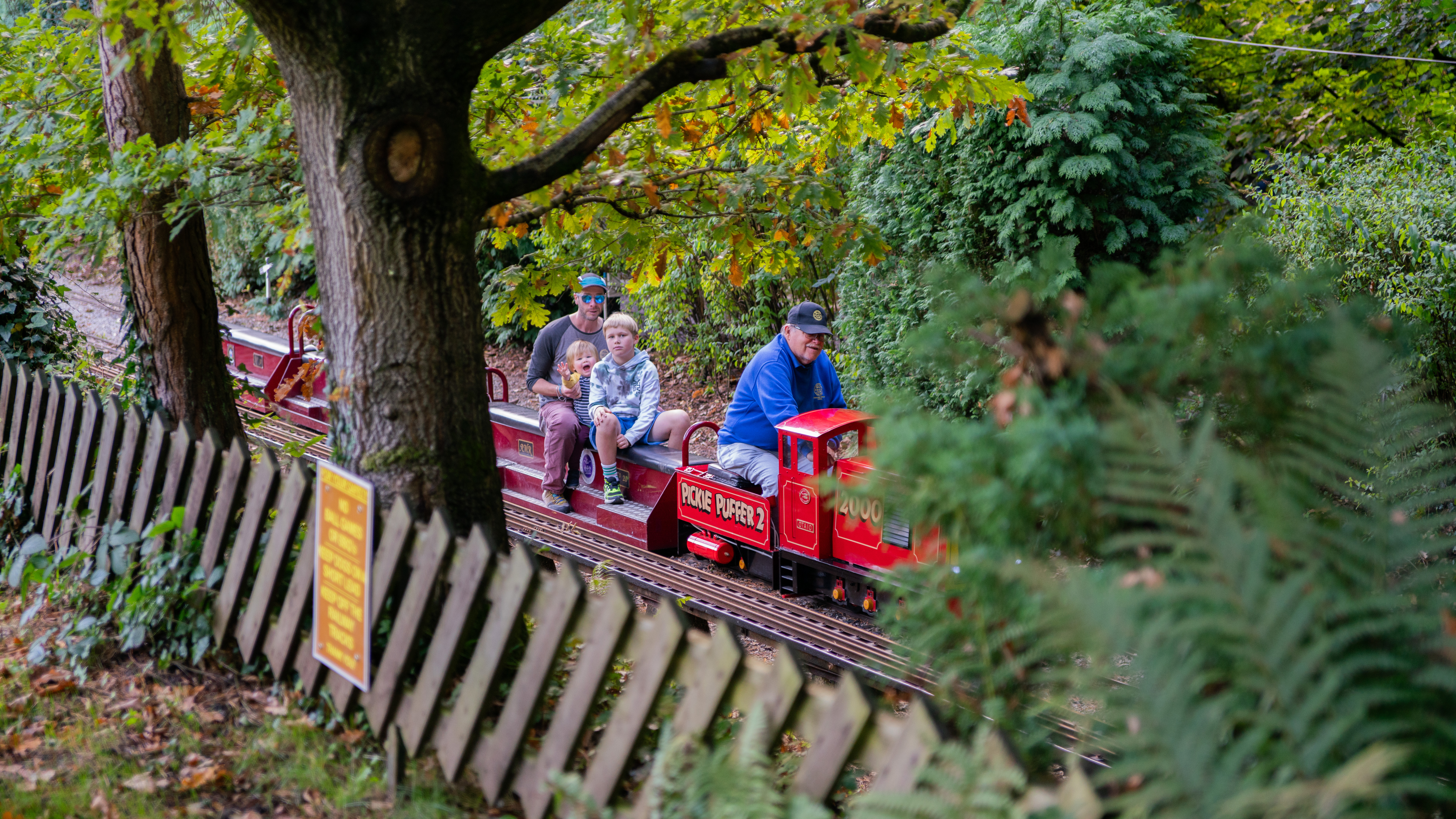 A small red miniature train travels along a track surrounded by trees and greenery. Several passengers are seated on open carriages behind the engine, passing a wooden fence and a yellow sign in a leafy outdoor setting.